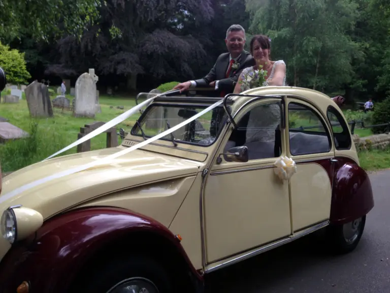 Bride and groom standing up through the open roof of a cream and maroon Citroën 2CV in wedding ribbons, in a churchyard