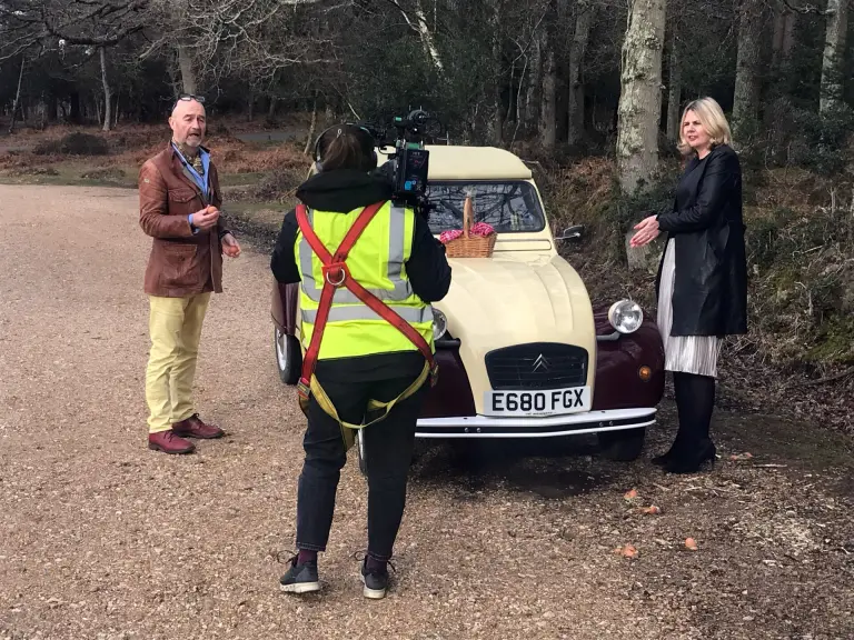 TV camera operator filming a man and woman beside Citroën 2CV E680 FGX in woodland, a wicker basket on its bonnet