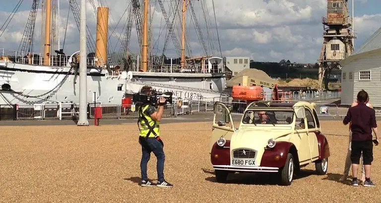 Cream and maroon Citroën 2CV E680 FGX being filmed by a TV camera operator on a quayside beside the historic ship HMS Gannet