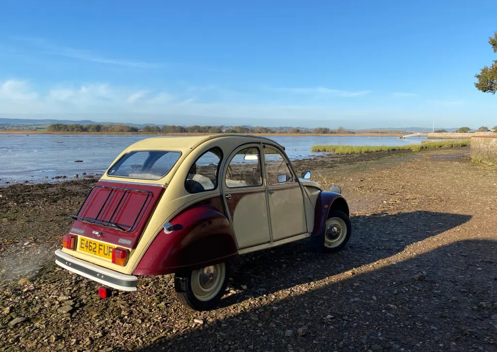 Cream and maroon Citroën 2CV E462 FUF parked on a shingle foreshore beside a calm estuary under a clear blue sky