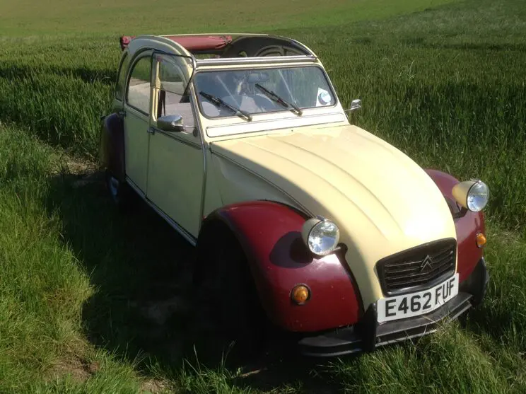 Cream and maroon Citroën 2CV E462 FUF with its roof rolled back, parked in a green crop field on a sunny day