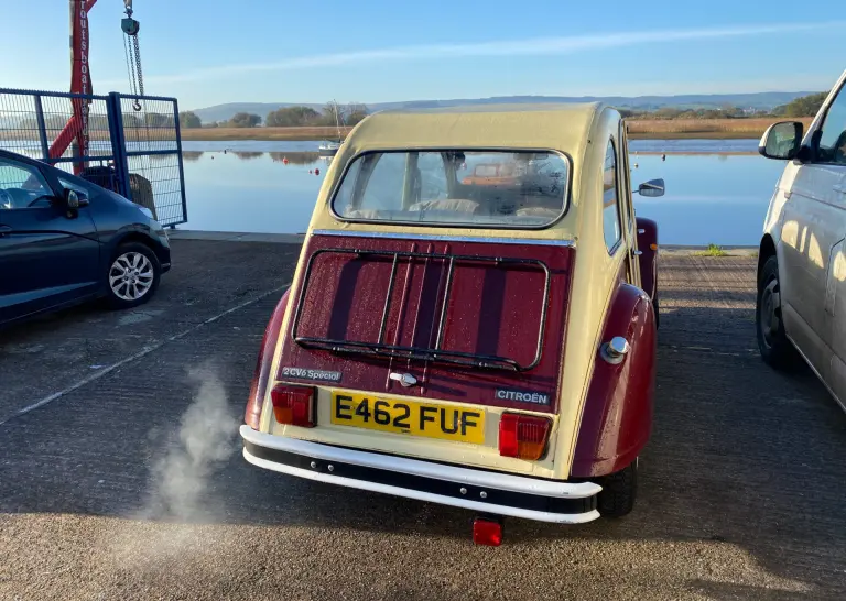 Rear of cream and maroon Citroën 2CV E462 FUF on a boatyard slipway beside calm water, exhaust vapour rising in low sun