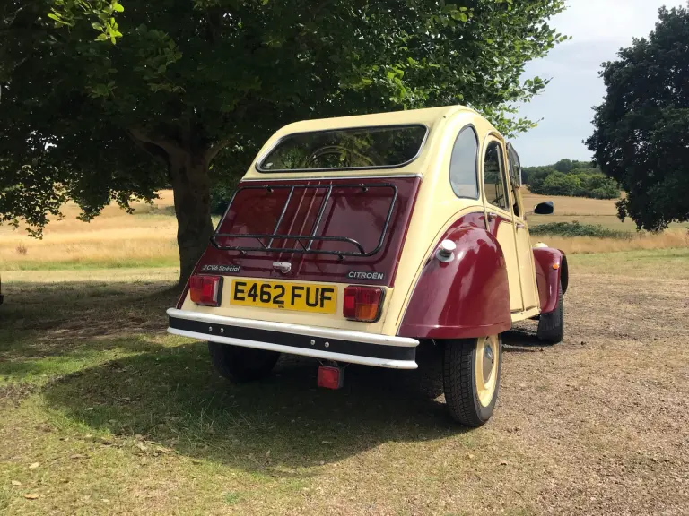 Rear view of cream and maroon Citroën 2CV E462 FUF with a luggage rack, parked in the shade of a tree in parkland