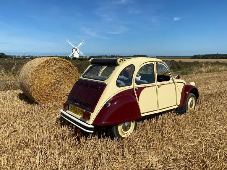Cream and maroon Citroën 2CV E462 FUF beside a round straw bale in a stubble field, a white windmill under blue sky beyond