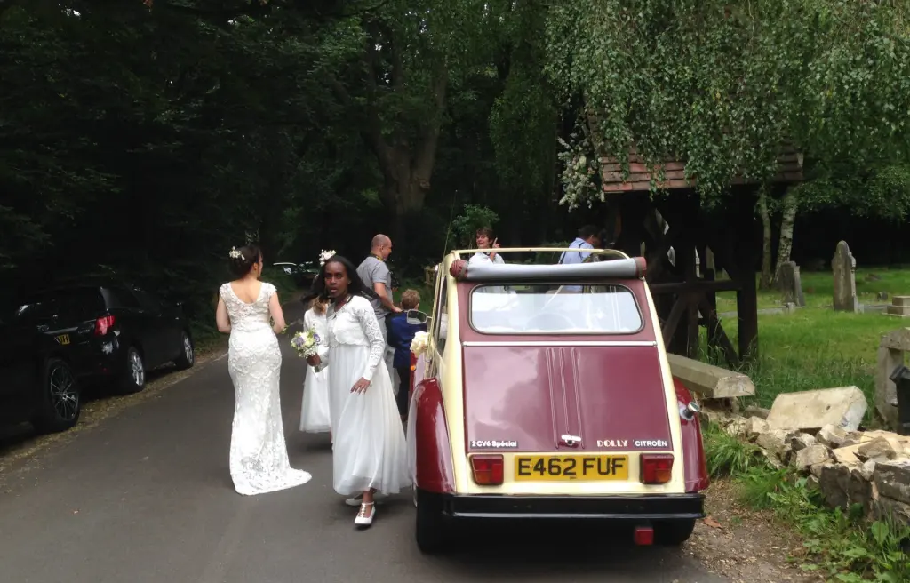 Rear of cream and maroon Citroën 2CV E462 FUF by a churchyard lychgate as the bride and bridesmaids walk along the lane