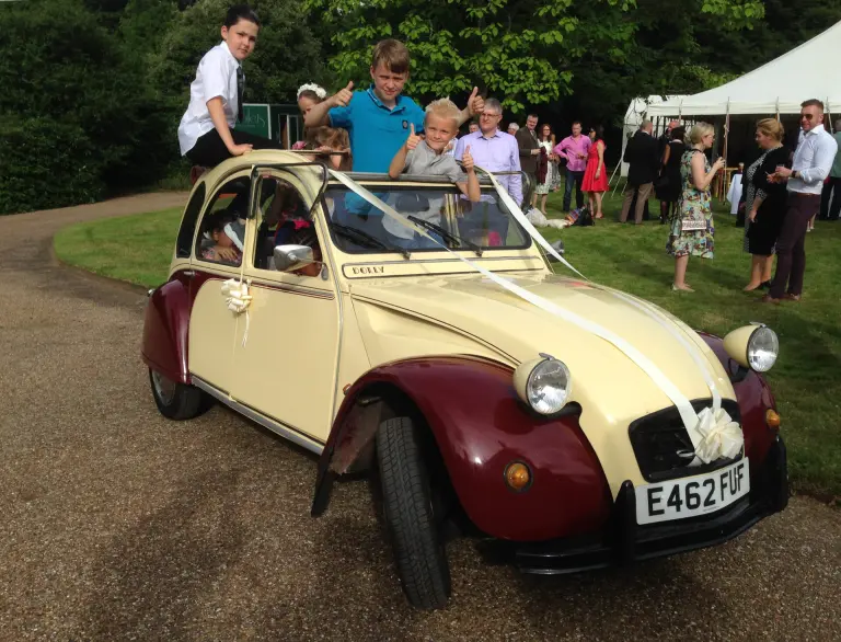Children giving thumbs up through the open roof of Citroën 2CV E462 FUF in wedding ribbons, guests by a marquee behind