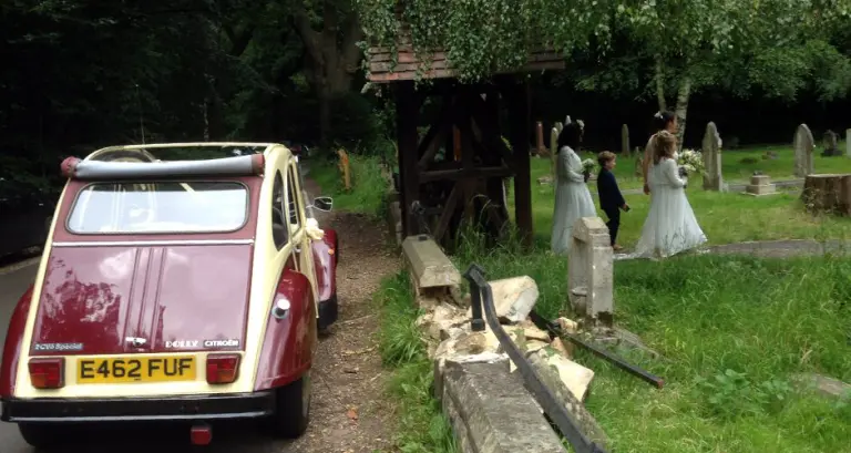 Rear of cream and maroon Citroën 2CV E462 FUF by a churchyard lychgate as bridesmaids and a page boy walk among the graves