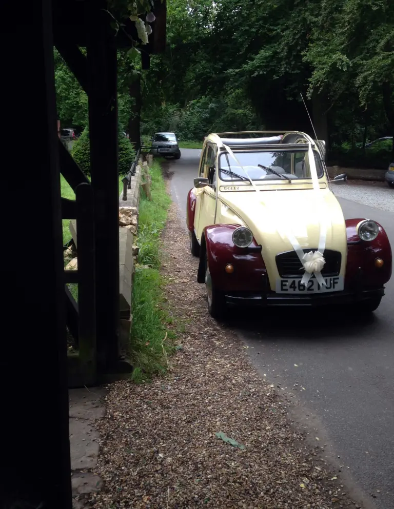 Cream and maroon Citroën 2CV E462 FUF in white wedding ribbons, parked on a tree-lined lane by a church lychgate