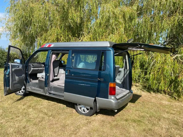 Teal and silver Mazda Bongo campervan with its doors and tailgate open, parked on grass beneath a weeping willow
