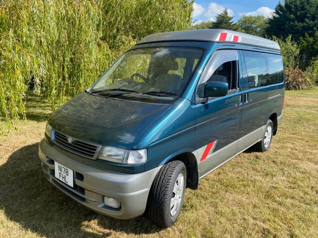 Teal and silver Mazda Bongo campervan, roof down, parked on grass beside a weeping willow on a sunny day