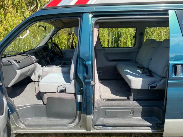 Interior of a teal Mazda Bongo seen through its open side doors, showing the cab, grey front seat and rear bench seat