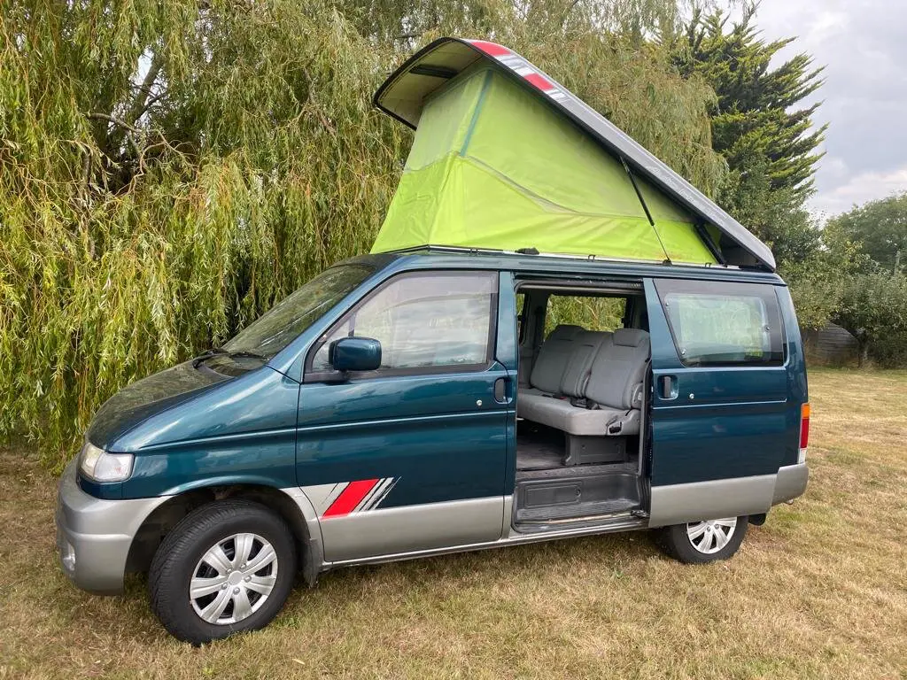 Teal and silver Mazda Bongo campervan with its pop-top roof raised to show green canvas sides, and its side door open