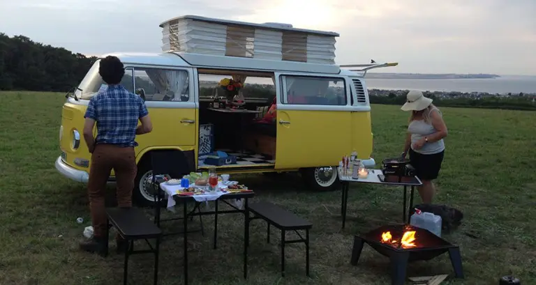 Yellow VW campervan with its roof raised on a hilltop above the sea at dusk, two people preparing a meal by a fire pit