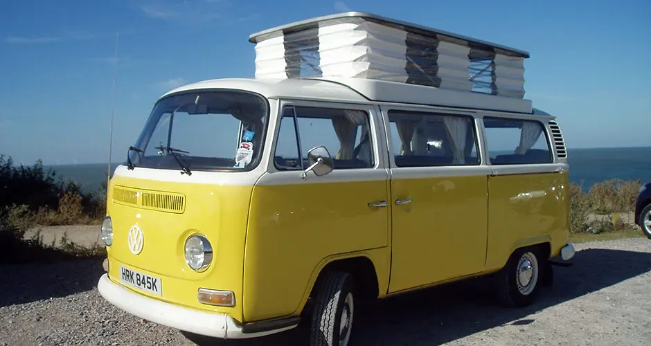 Yellow and white VW campervan with its pop-top roof raised, parked on gravel above the sea under a blue sky