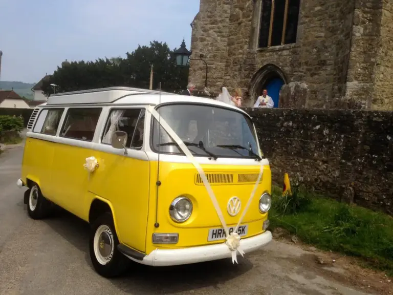 Yellow and white VW campervan with white wedding ribbons and bows outside a stone church, the bride and vicar at its door